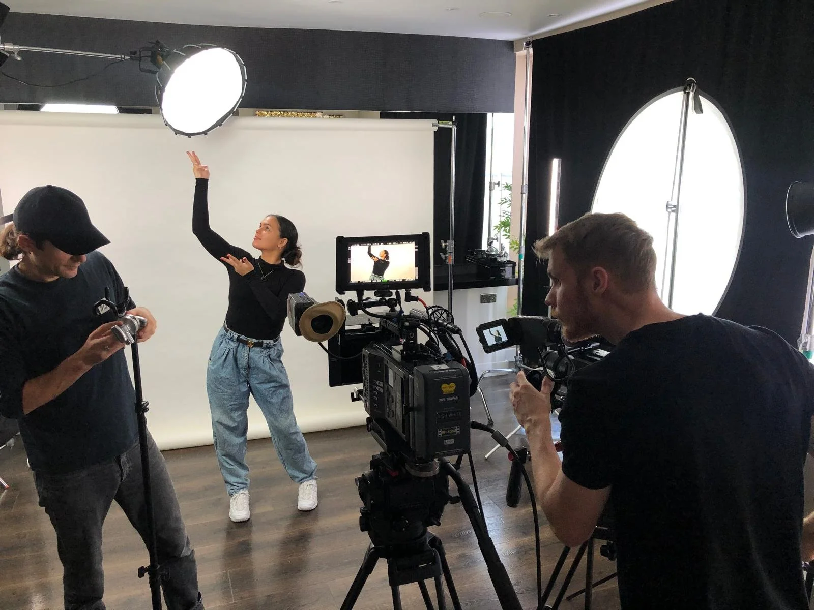 A woman poses with her arms raised and fingers forming a peace sign during a video shoot, with two crew members adjusting equipment and a camera operator filming her in front of a white backdrop, inside a studio with black walls and large circular light diffusers.