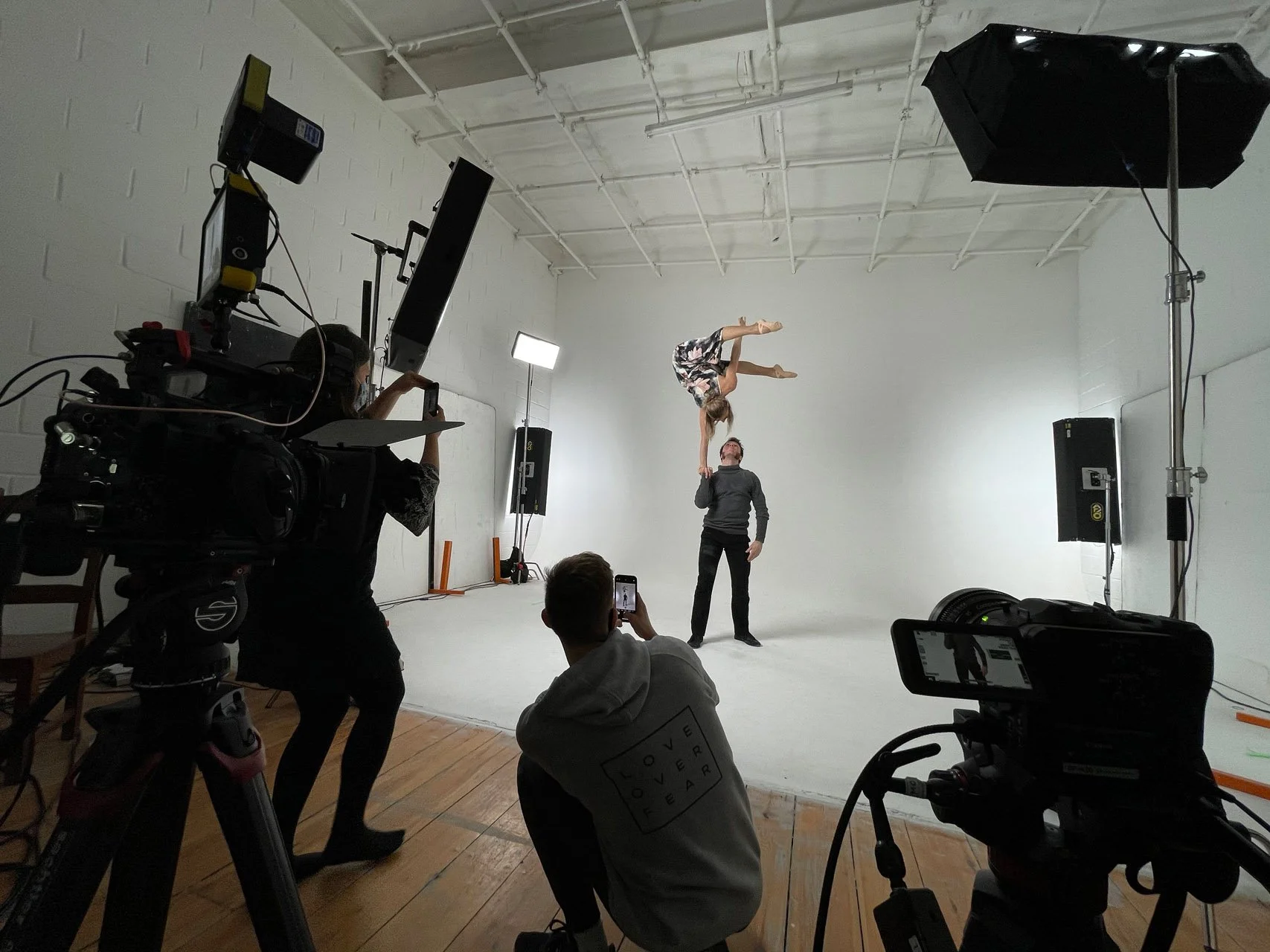 A dancer balancing on a person's hand in a studio with group of people filming and photographing.