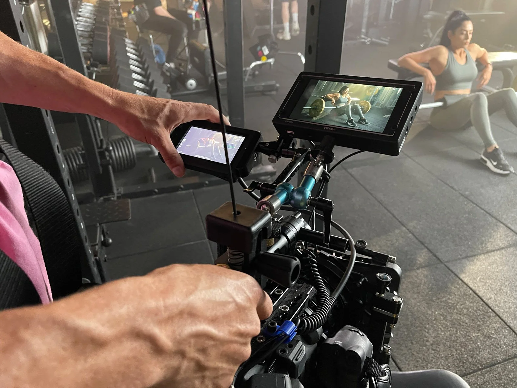 A person operating a camera rig captures a woman doing a seated stretch exercise on a bench in a gym. The woman is wearing a black sports bra and leggings, with her hands on her hips, and appears focused.
