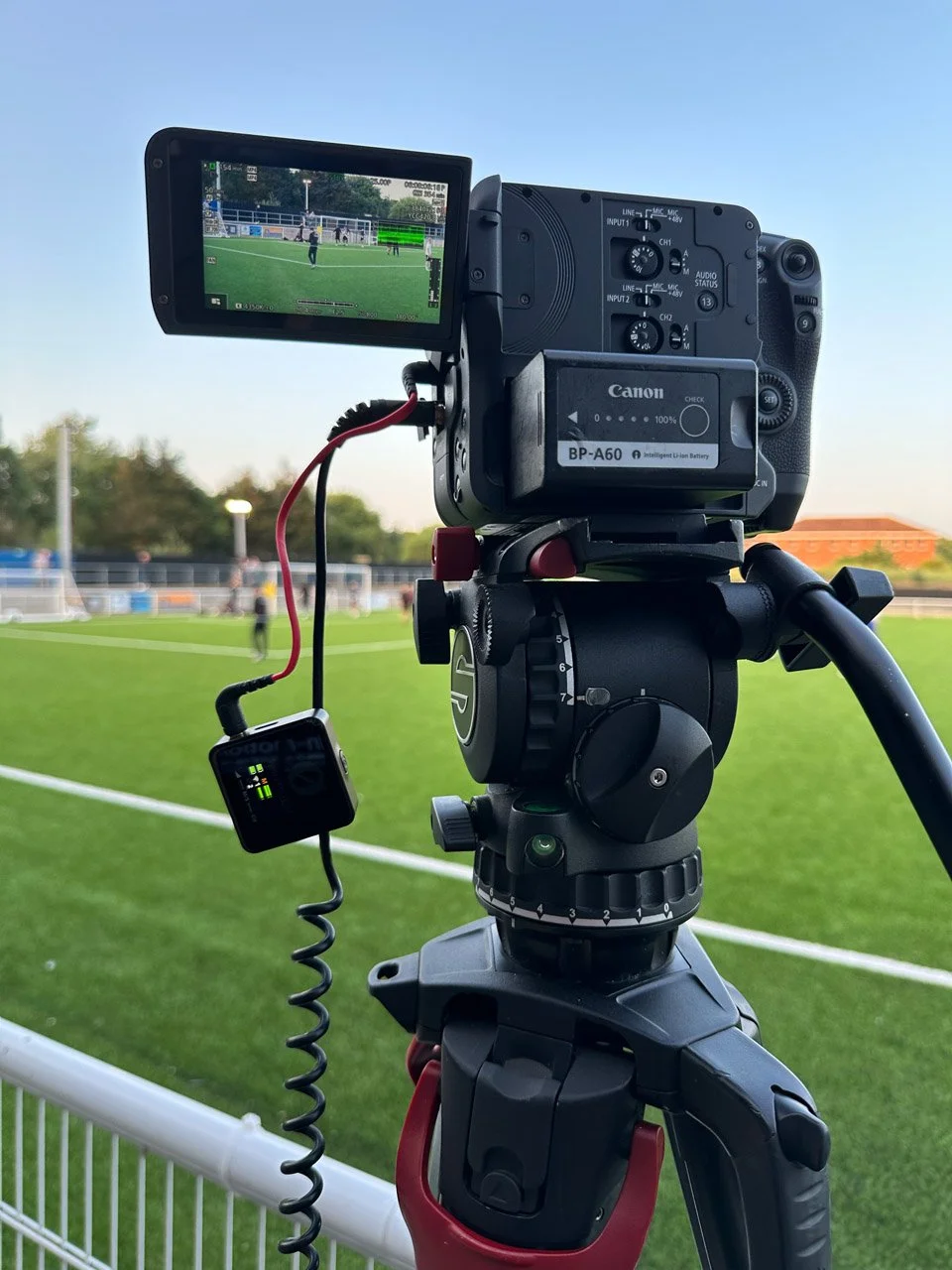 Professional video camera mounted on a tripod at a soccer field during daytime, with the camera view showing players on the field.