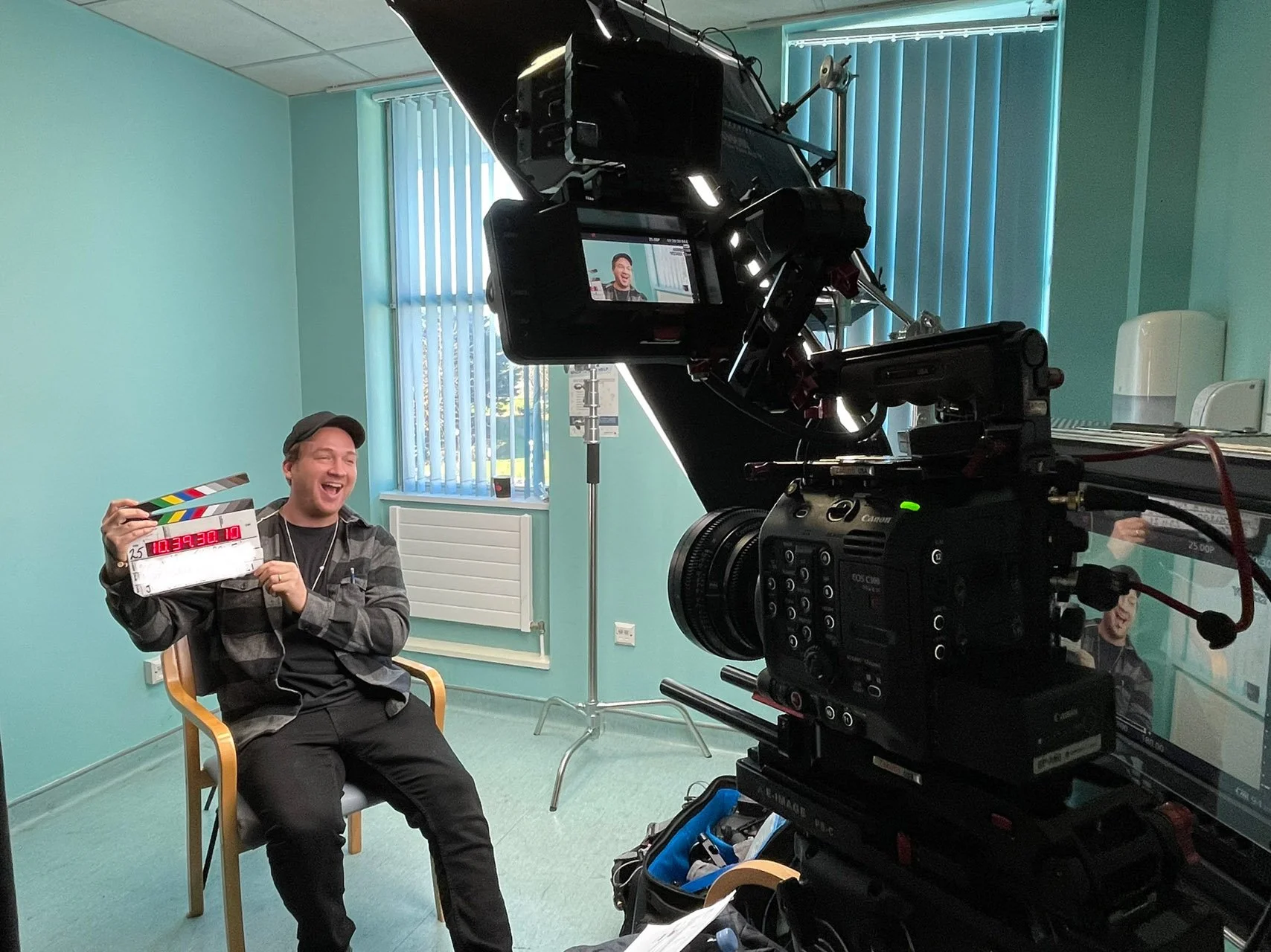 A man sitting in a chair holding a clapperboard, appearing to smile and laugh during a video or film shoot in a room with teal walls and vertical blinds.