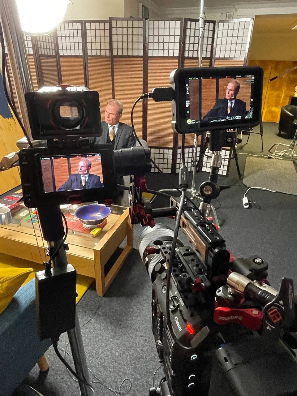 A recording or interview setup with two cameras focused on a man in a suit, seated near a wooden table in front of a partition. The scene includes professional lighting and various equipment, indicating a video production or interview in progress.