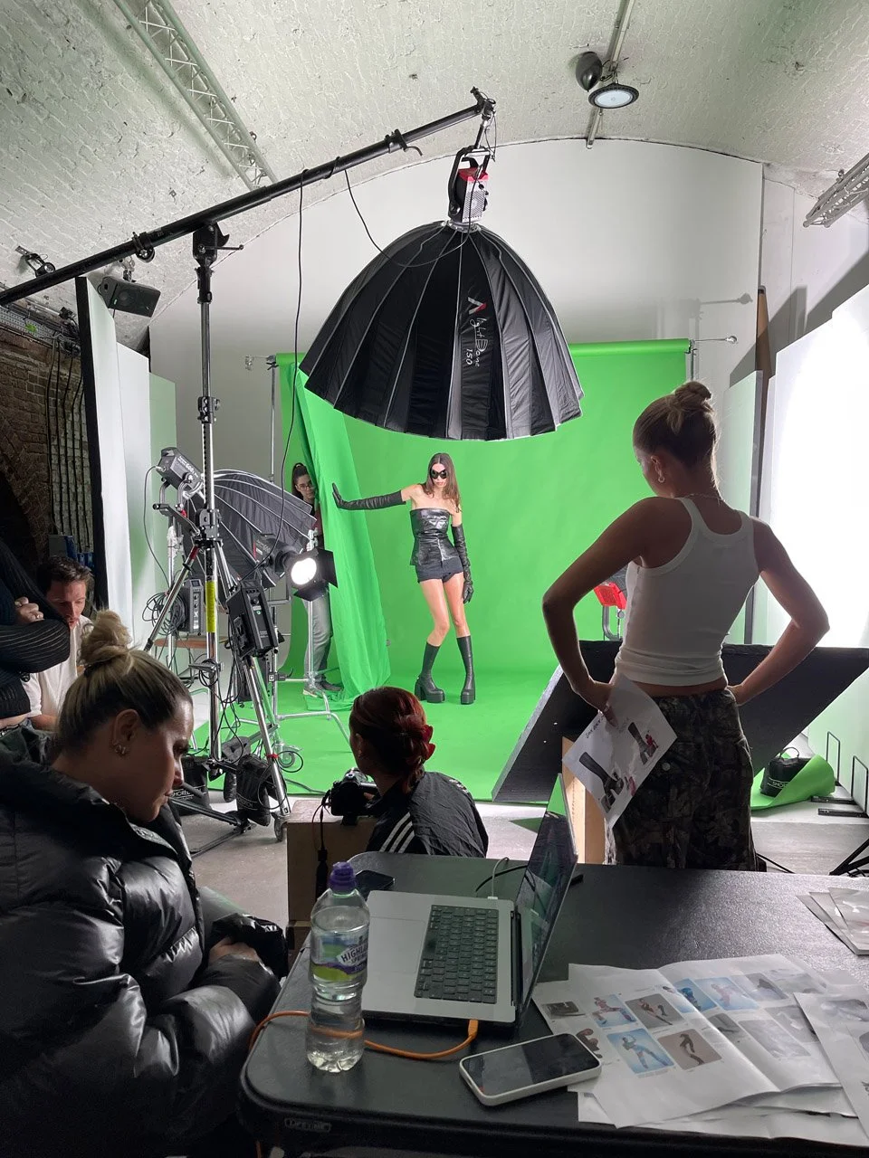 Models pose in a photography studio with a green screen background, photography equipment, and crew members preparing for a photoshoot.