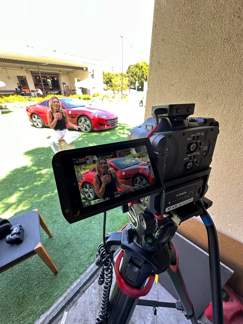 A video camera on a tripod capturing a woman in black top and white pants standing next to a red sports car outside a building.