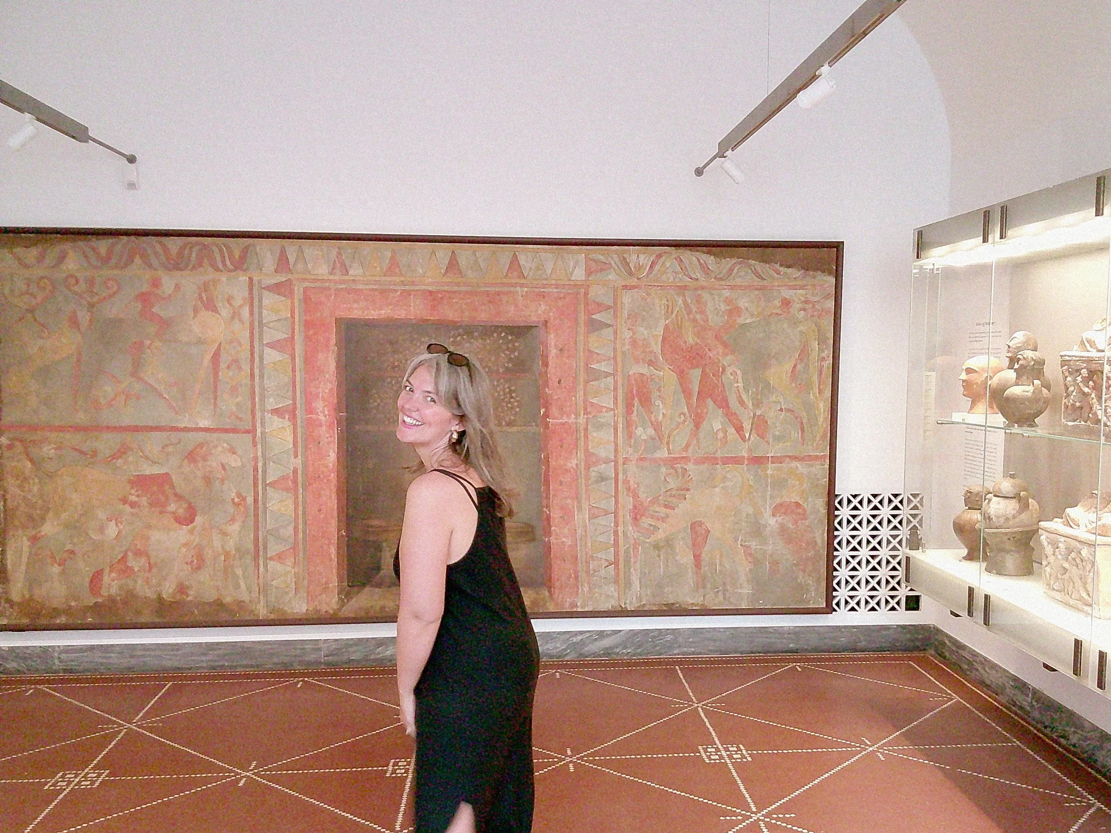 A woman in a black dress standing in front of an ancient mural painting in a museum, surrounded by display cases with artifacts.