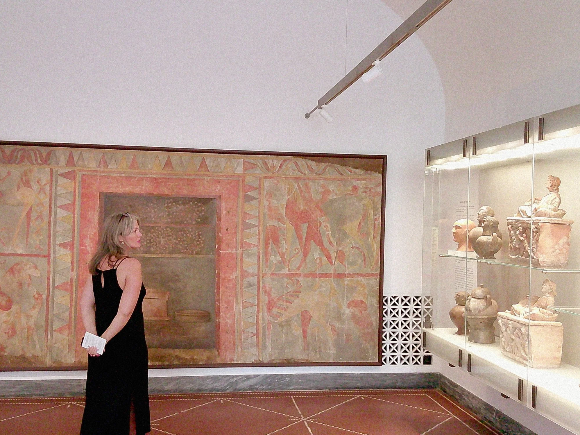 A woman in a black dress holding a brochure looking at ancient artifacts in a museum display case.