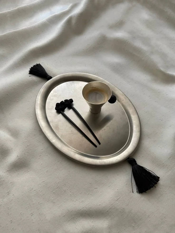 A round metal tray with black tassels on two sides, holding a small beige cup, black chopsticks, and a black spoon, placed on a white fabric surface.