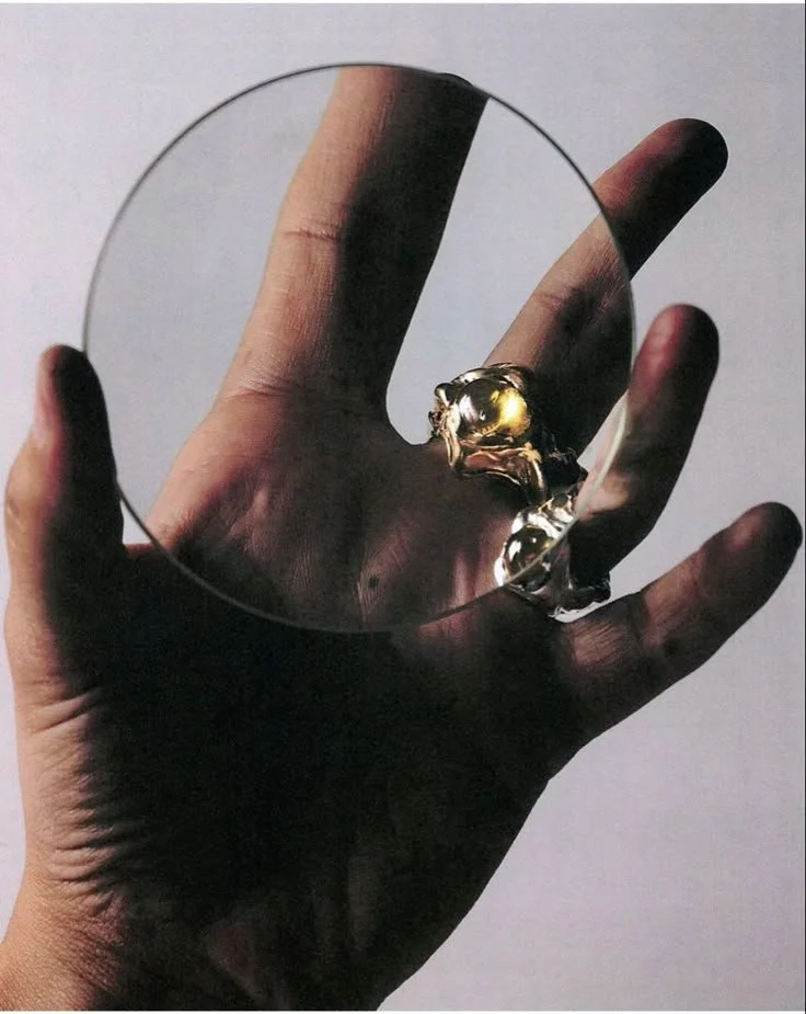 A person holding a round mirror with a golden ring reflection, showing their hand.