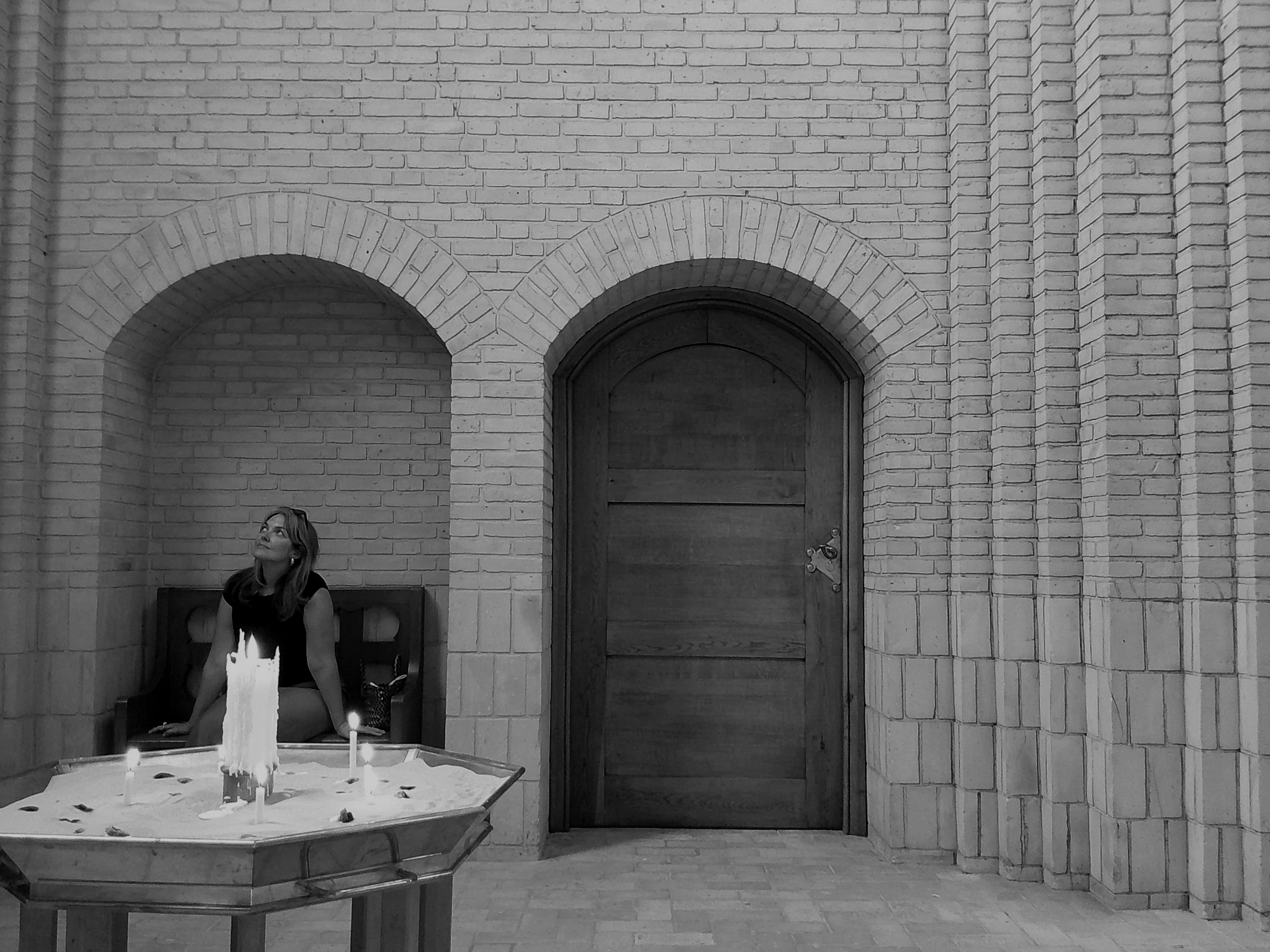 A woman sitting on a bench in front of a brick wall with an arched door, with candles and a cake on the table in front of her, and a candlelit celebration setup.