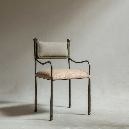 An antique metal-framed chair with beige cushioned seat and backrest, set against a plain, light-colored background.