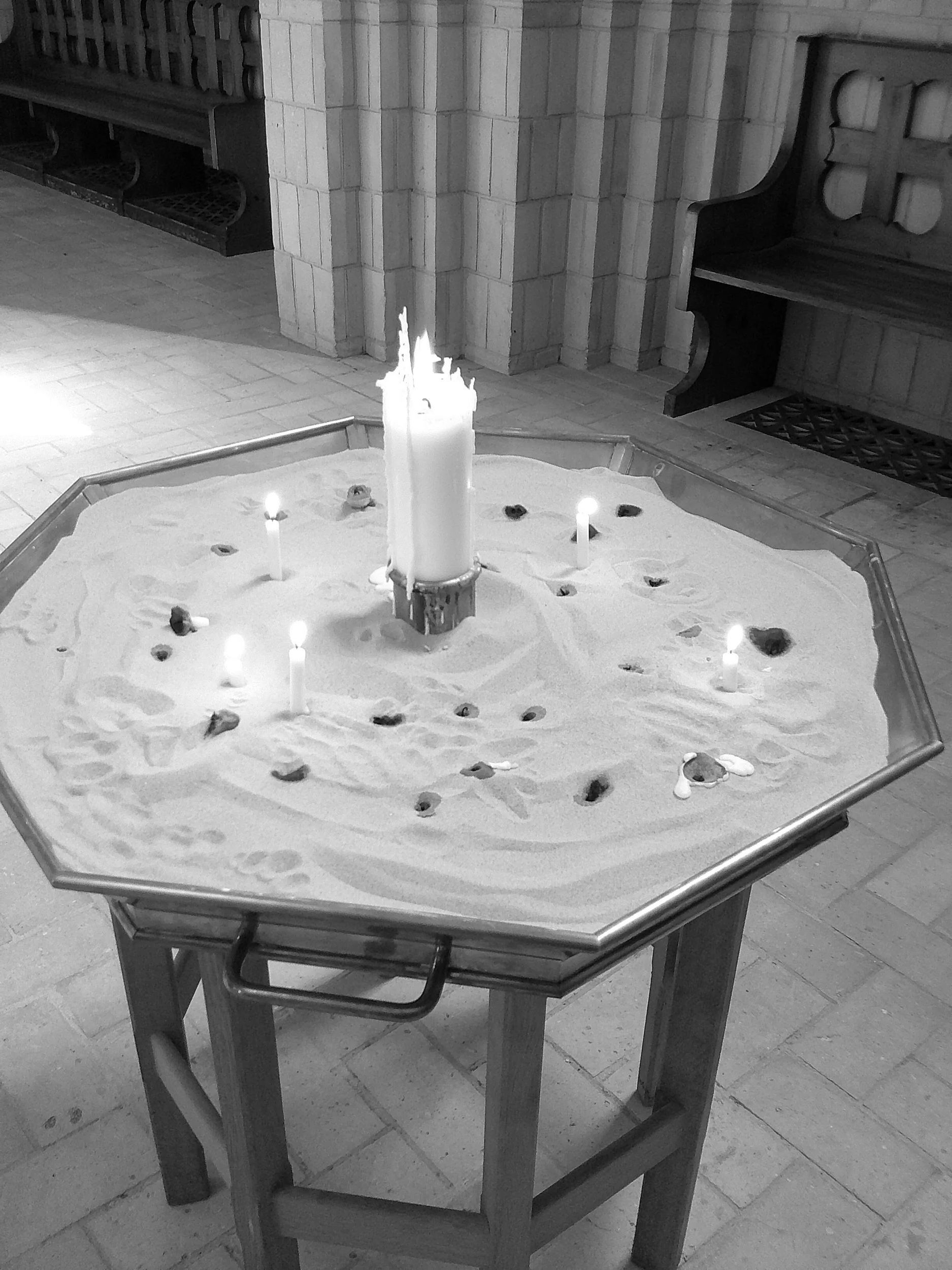 A church altar with a sand-covered surface, candles, and small stones, surrounded by wooden pews and stone walls.