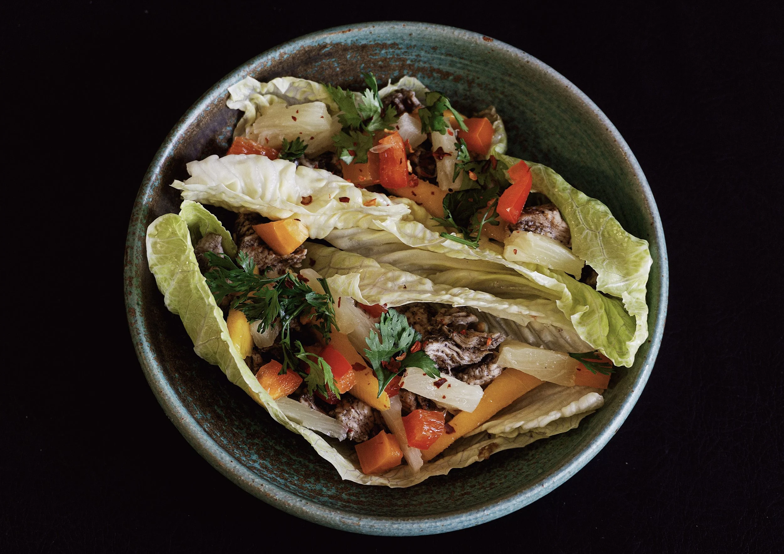 Jerk chicken lettuce wraps resting in a handmade ceramic bowl topped with chopped pineapple, chopped bell peppers, cilantro and red pepper flakes.