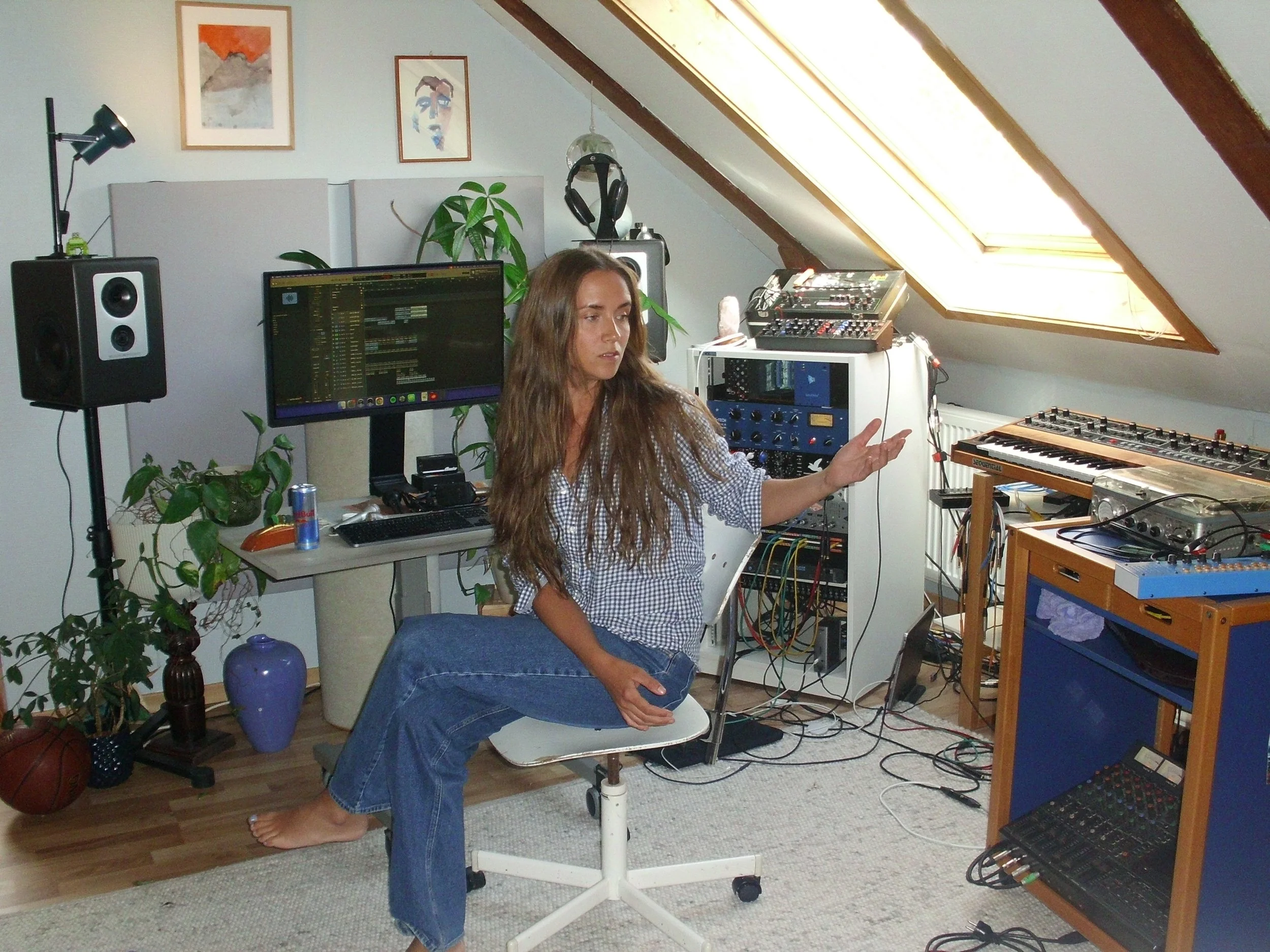 A woman with long brown hair, wearing a checkered shirt and jeans, is sitting barefoot on a white chair in a room with sloped ceilings and a skylight. She appears to be talking or explaining something, with her right hand raised. The room is filled with musical equipment, including speakers, a computer monitor, synthesizers, and audio gear. There are plants, artwork on the wall, and a Red Bull energy drink can on the desk.