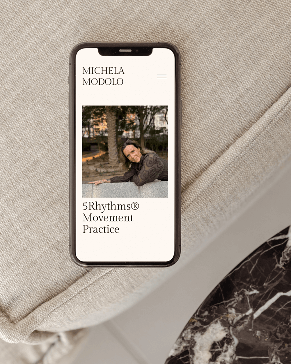 Smartphone displaying a website for Michela Modolo with a photo of a smiling woman leaning on a bench and text listing a movement practice called 5Rhythms Movement Practice.