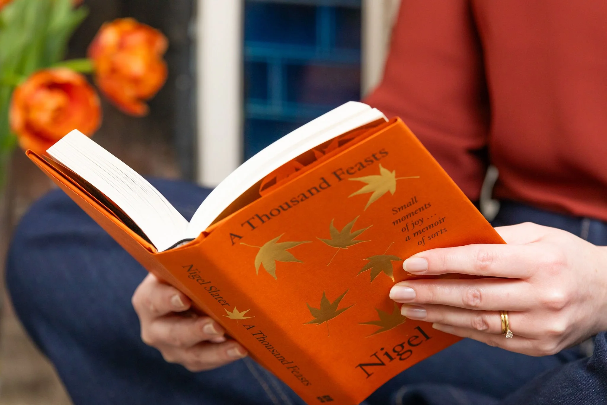 A person holding an orange book titled "A Thousand Forests" by Nigel Slater, with an orange and green leaf design on the cover, sitting indoors near tulips.