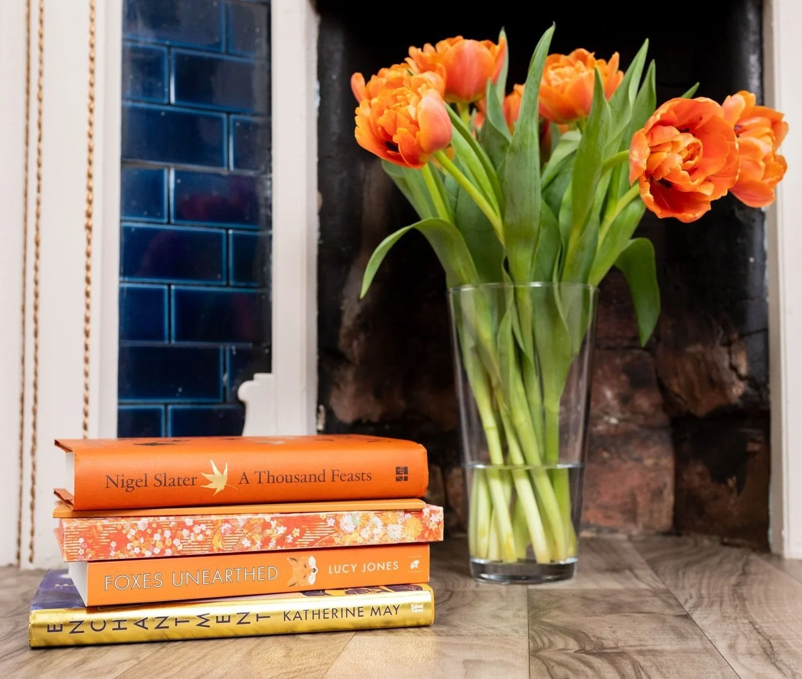 A stack of four books with orange and yellow covers next to a glass vase of orange tulips on a wooden surface, with a background of a dark brick wall and a white window frame.