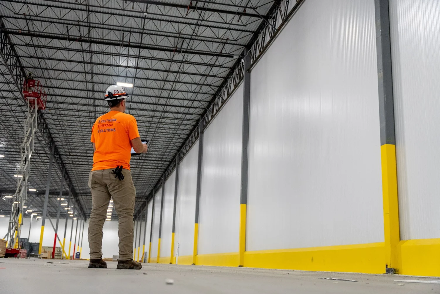 A worker wearing an orange shirt with the text 'Southern Thermal Solutions', tan pants, and a white safety helmet stands inside a large industrial warehouse. The worker is looking at a tablet device, and the warehouse features high ceilings with metal beams and walls with yellow safety guards along the bottom.