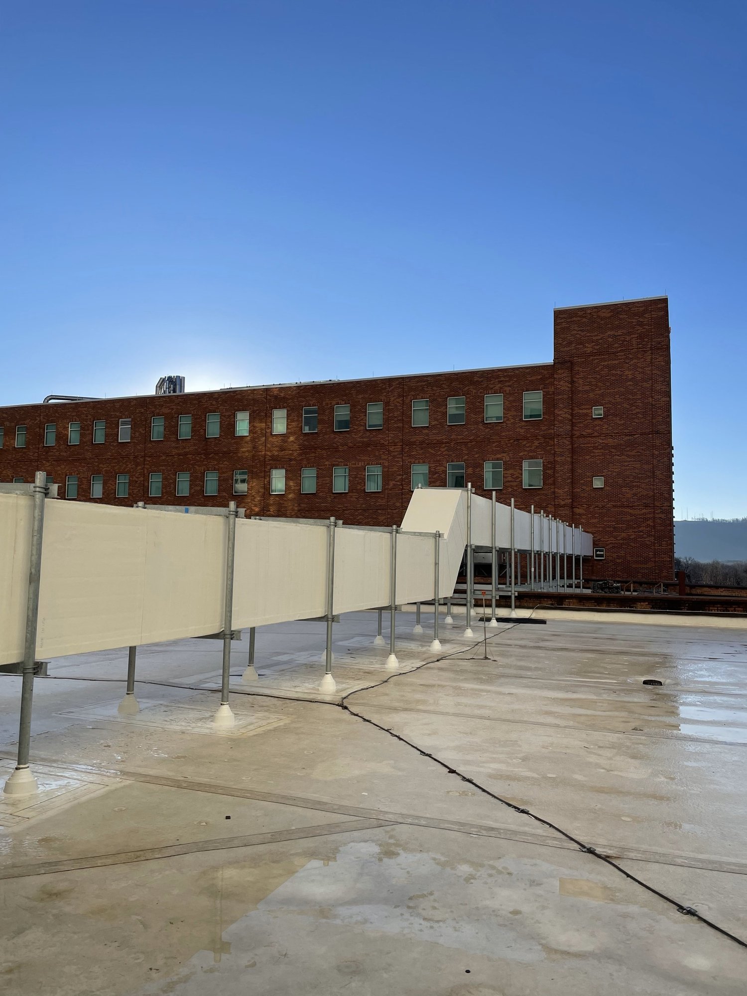 View of a rooftop with a large brick building in the background, an HVAC duct running across the rooftop, and a clear blue sky.
