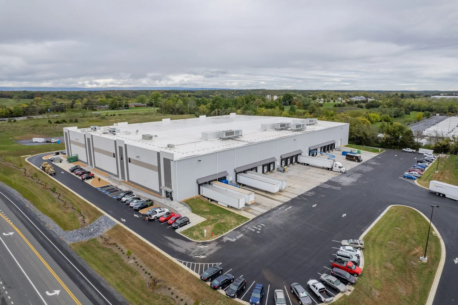 An aerial view of a large warehouse with multiple loading docks and trucks, surrounded by parking lots filled with cars. The warehouse is situated near a highway with a landscaped area and green fields in the background.