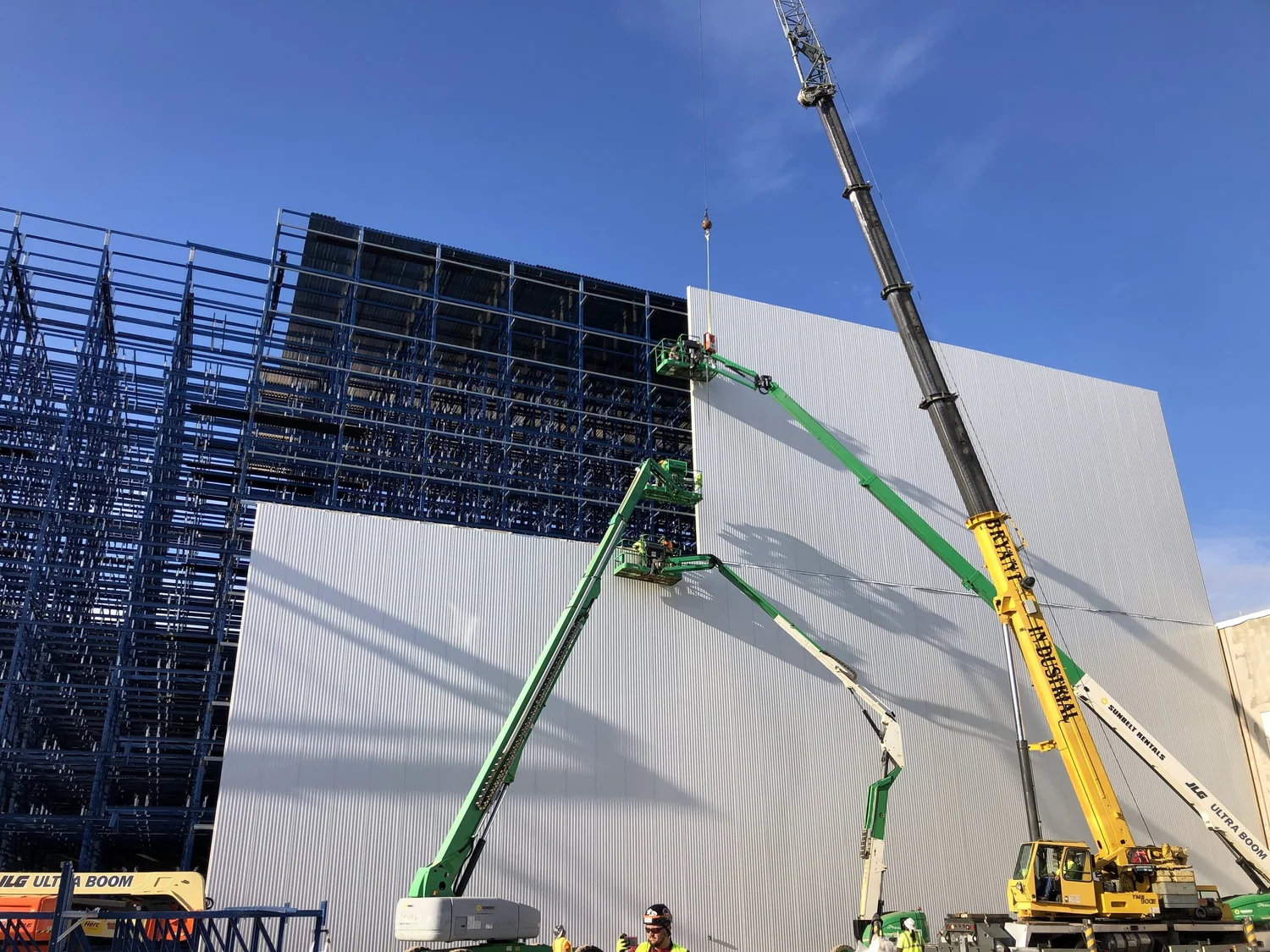 Construction workers using green and white boom lifts to work on a large white wall of a building, while a crane positions equipment against the structure. The building's exterior features a grid-like, dark-colored framework.
