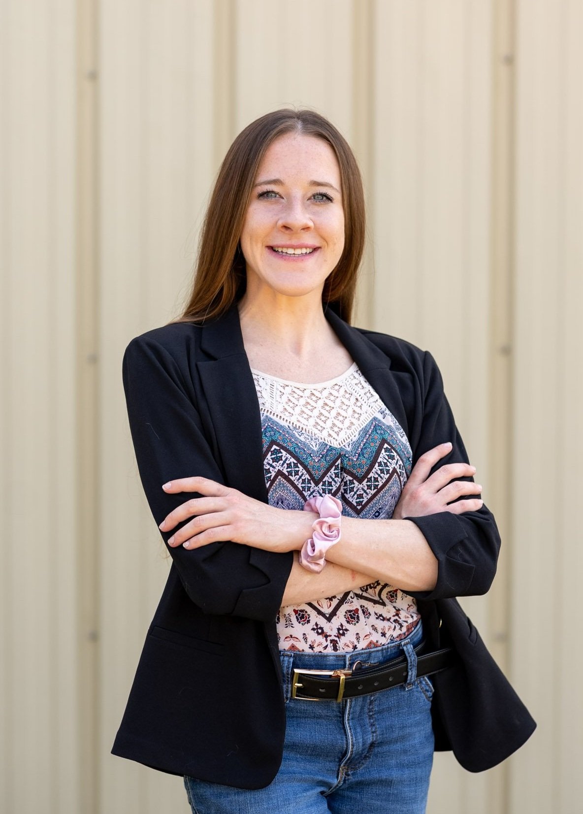A woman with long brown hair, wearing a black blazer over a patterned top, crossing her arms and smiling in front of a beige wooden background.