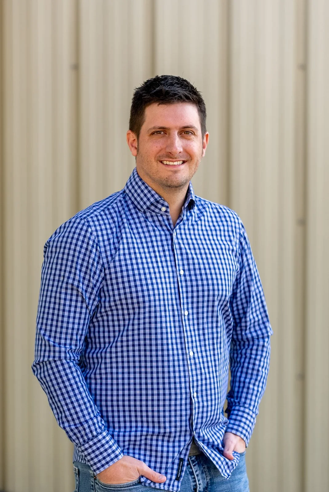 A man with short dark hair and a beard, wearing a blue and white checkered shirt, standing in front of a beige corrugated metal wall, smiling with his hands in his pockets.