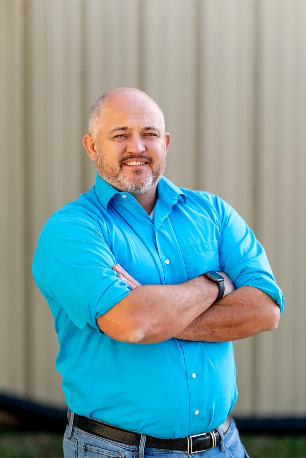 A man with a beard and short hair, wearing a bright blue button-up shirt and a smartwatch, standing with arms crossed in front of a beige background, smiling at the camera.