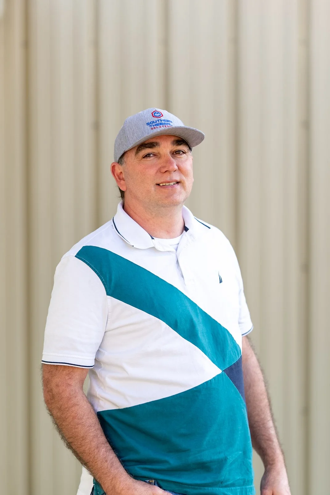 A man wearing a gray baseball cap with a logo and a white polo shirt with blue and teal geometric patterns, standing in front of a beige curtain.