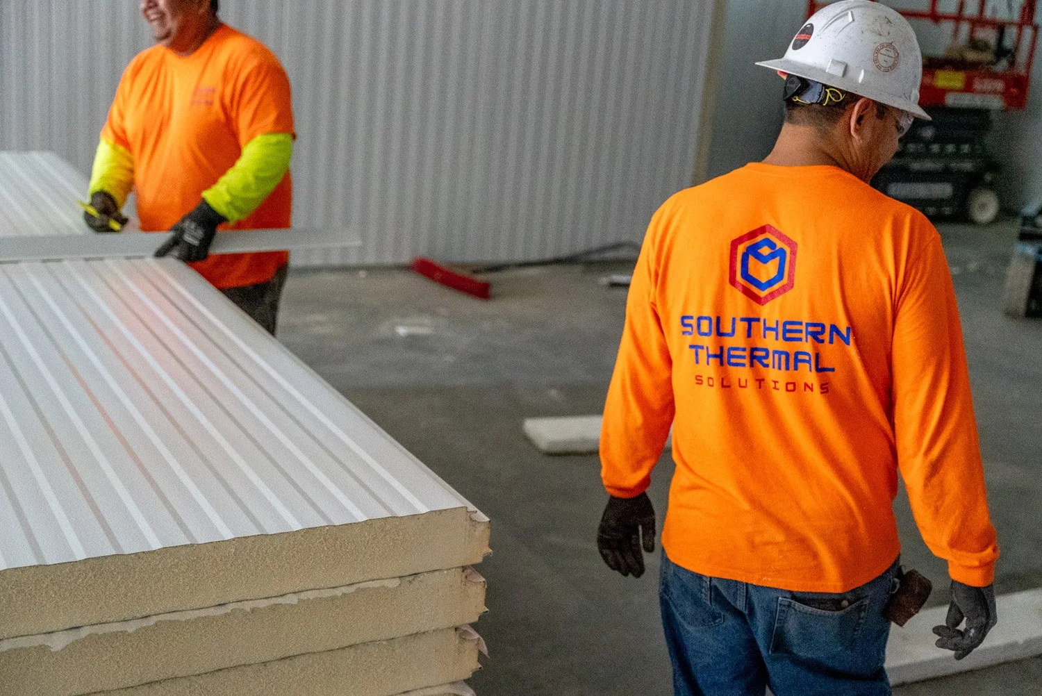 Construction workers in orange shirts and white helmets moving insulated panels in a warehouse or workshop.