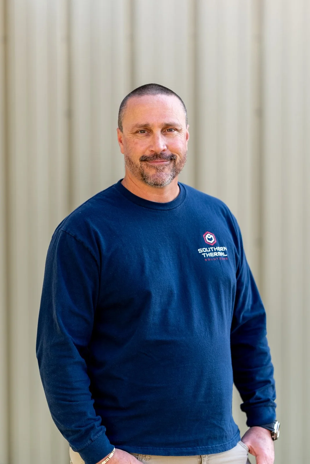 A man with short dark hair, beard, and mustache wearing a navy blue long-sleeve shirt with a logo that says "Southern Thermal Solutions" standing in front of a light-colored curtain.