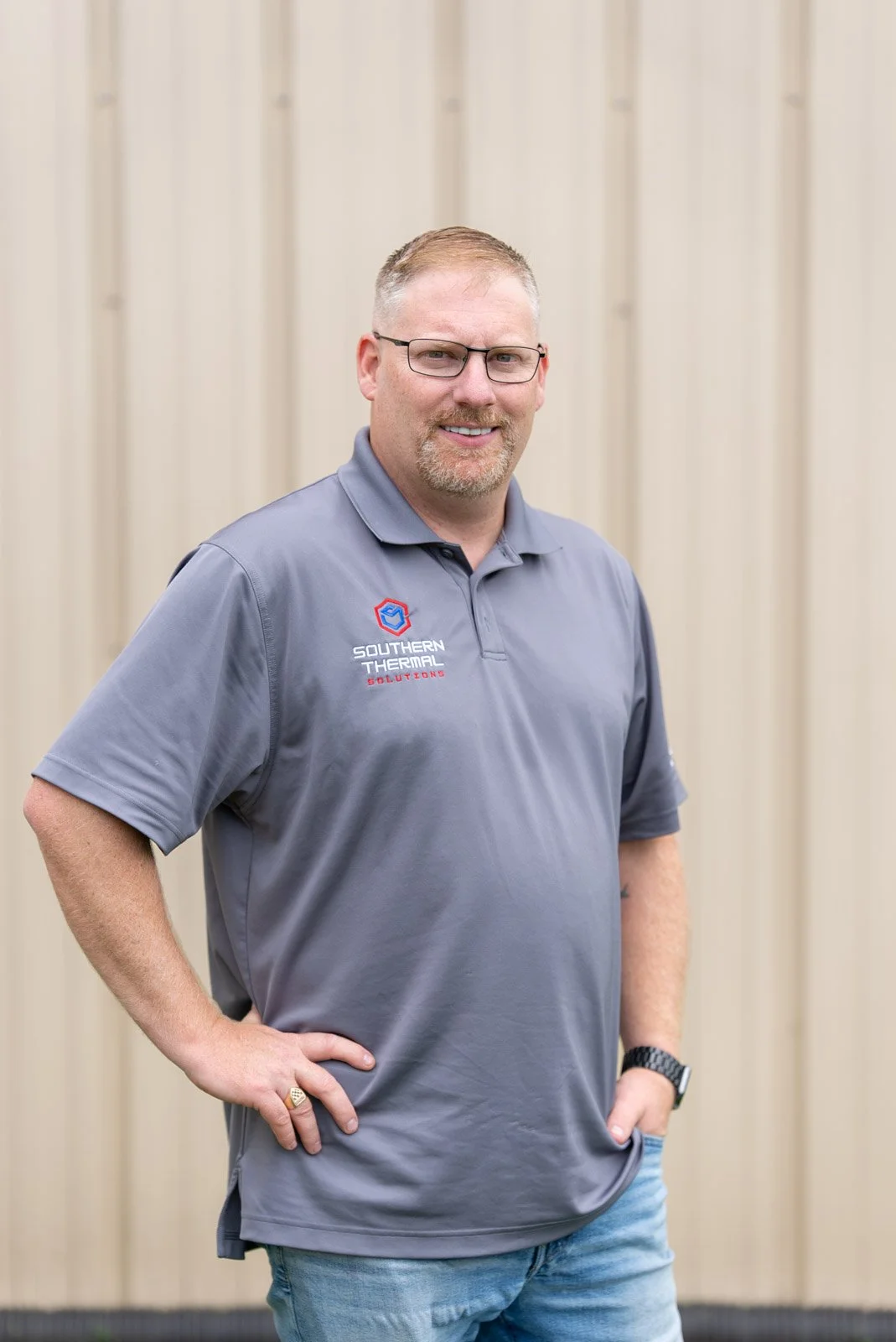 A man with glasses, a beard, and short hair, wearing a gray polo shirt with a logo that reads 'Southern Thermal Solutions,' stands in front of a beige wall, smiling with his hands on his hips.