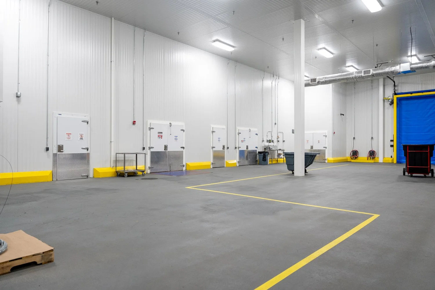 Empty industrial warehouse with gray concrete floor, white metal walls, and a gray ceiling with fluorescent lighting. There are several white metal doors with warning signs along the wall, yellow safety curbs, a cart, garbage bins, and a blue retractable door on the right side.