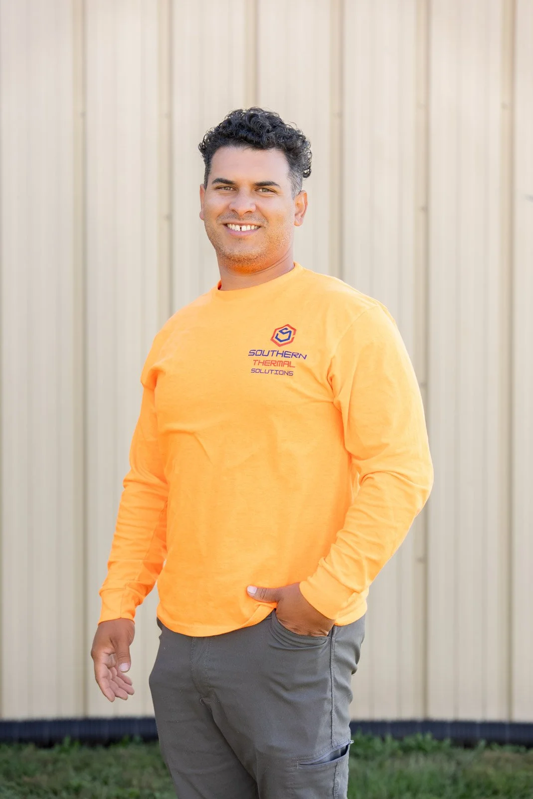 A man with curly dark hair, smiling, standing outdoors in front of a beige metal wall, wearing an orange long-sleeve shirt with a logo and text that reads 'Southern Thermal Solutions', and gray pants.