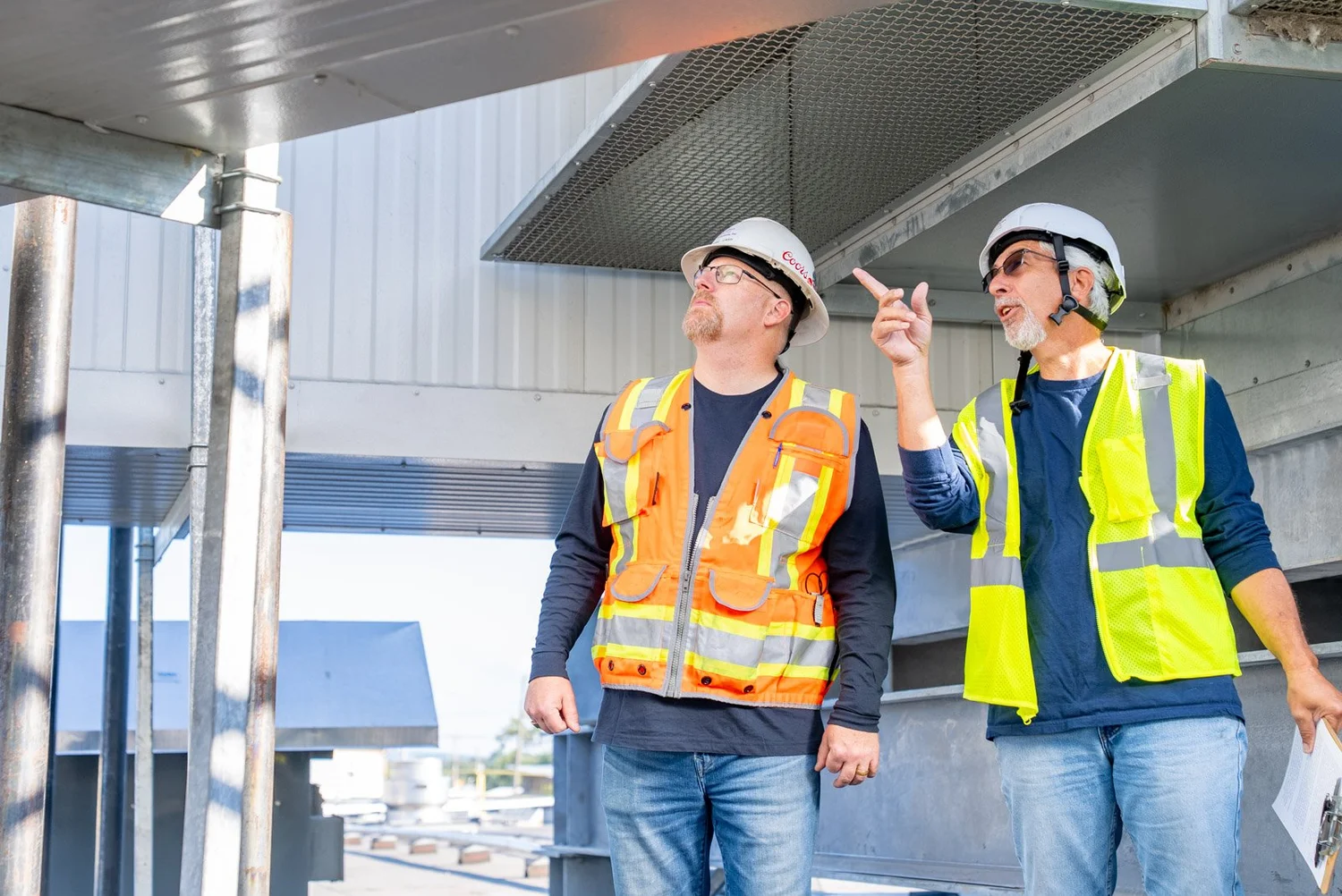 Two crew members in hard hats pointing out details under a rooftop duct system