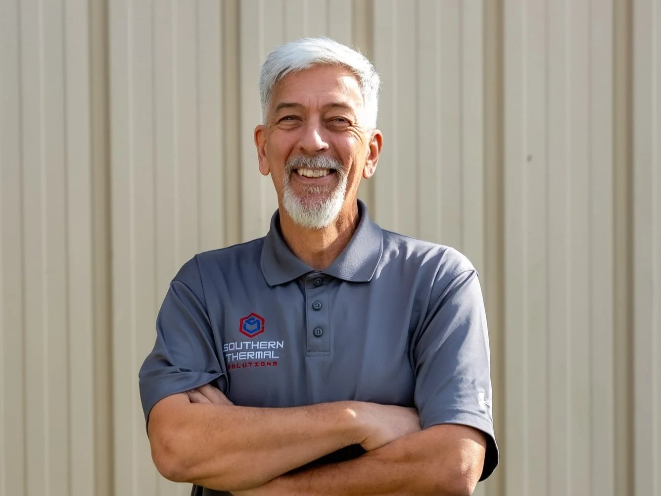 Smiling older man with gray hair and beard wearing a gray polo shirt with a company logo, standing outdoors with folded arms in front of a beige metal wall.