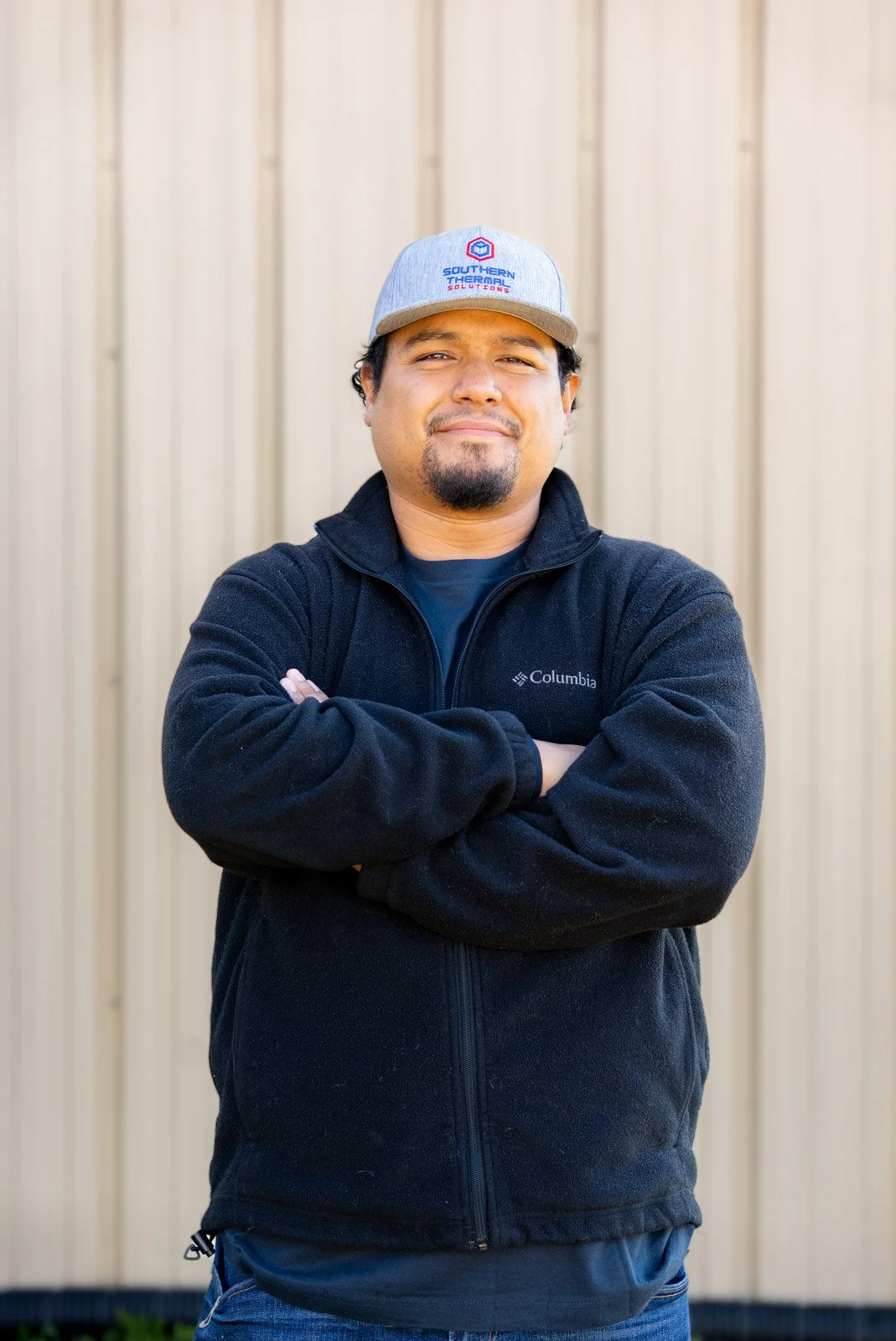 A man with crossed arms standing outdoors in front of a beige wooden wall, wearing a black Columbia fleece jacket and a gray baseball cap with a blue and red logo.