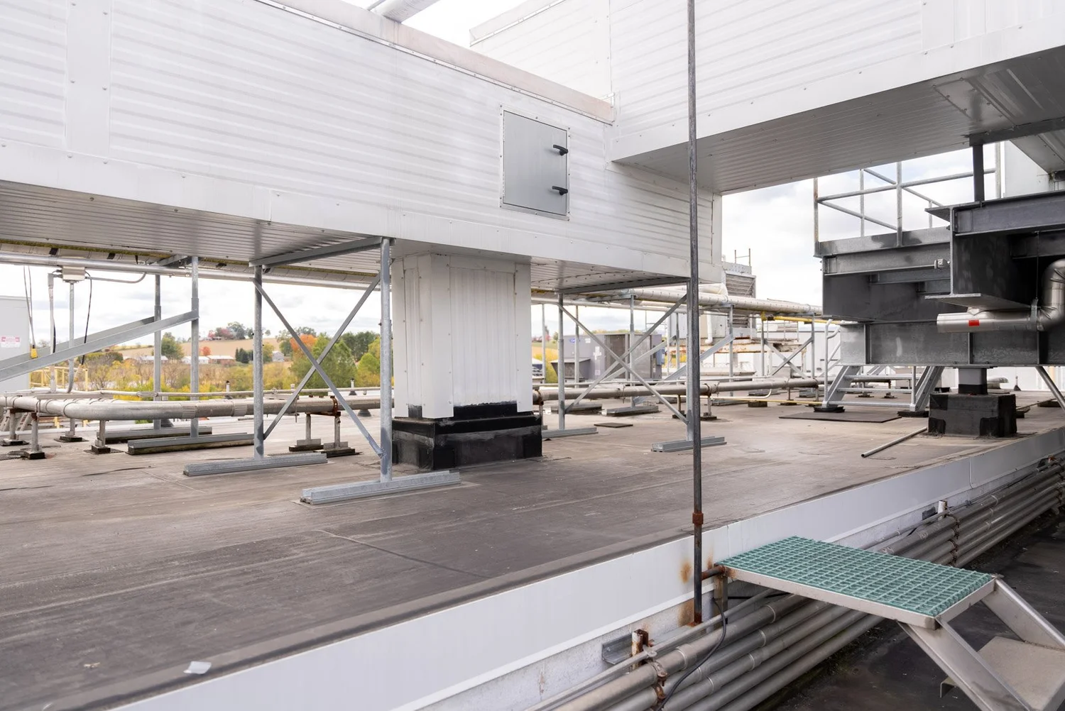 Rooftop of a building with industrial HVAC and piping, metal scaffolding, and a view of trees and sky in the distance.