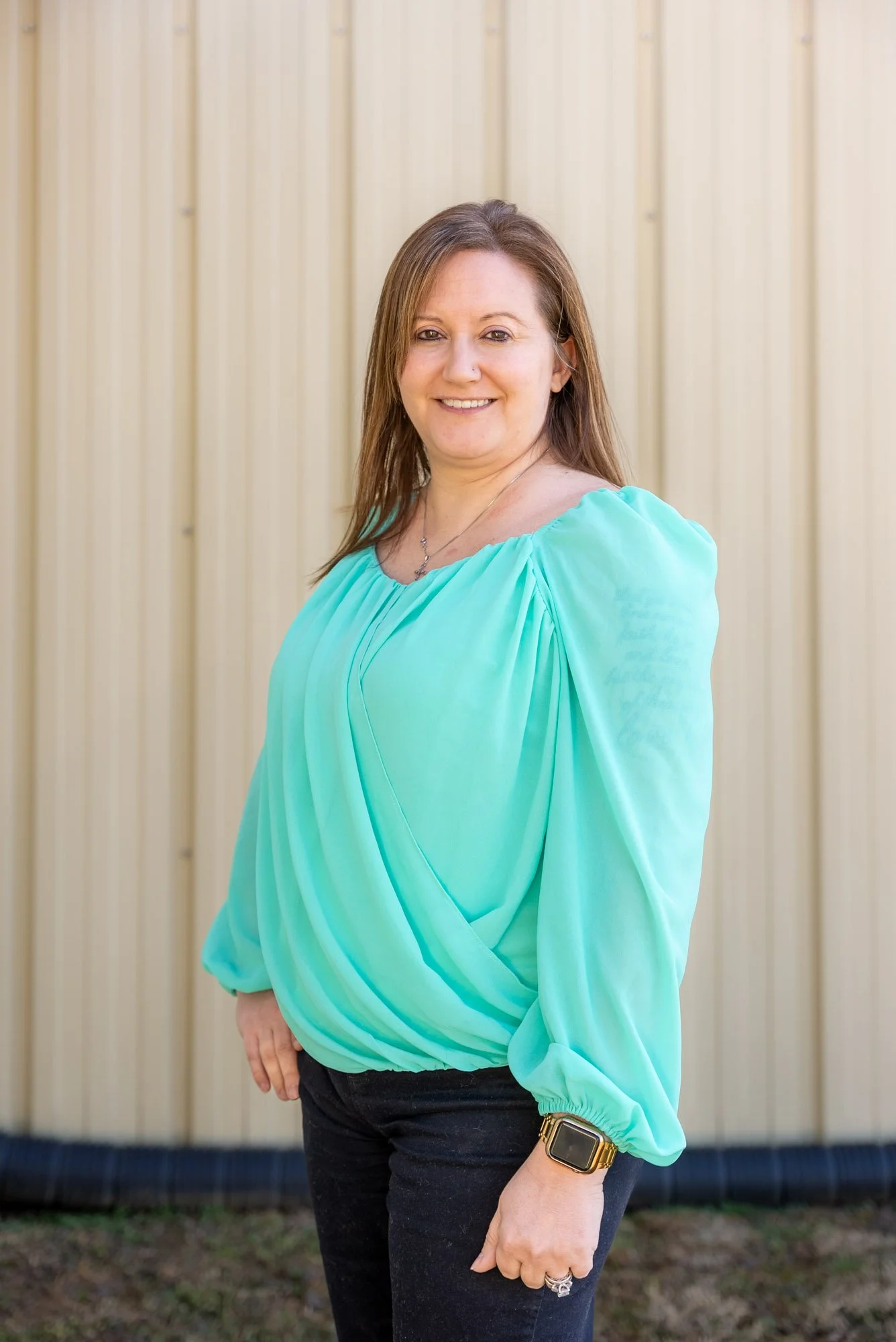 Woman with light brown hair wearing a long-sleeved turquoise blouse standing outdoors in front of a light-colored fence, smiling at the camera.