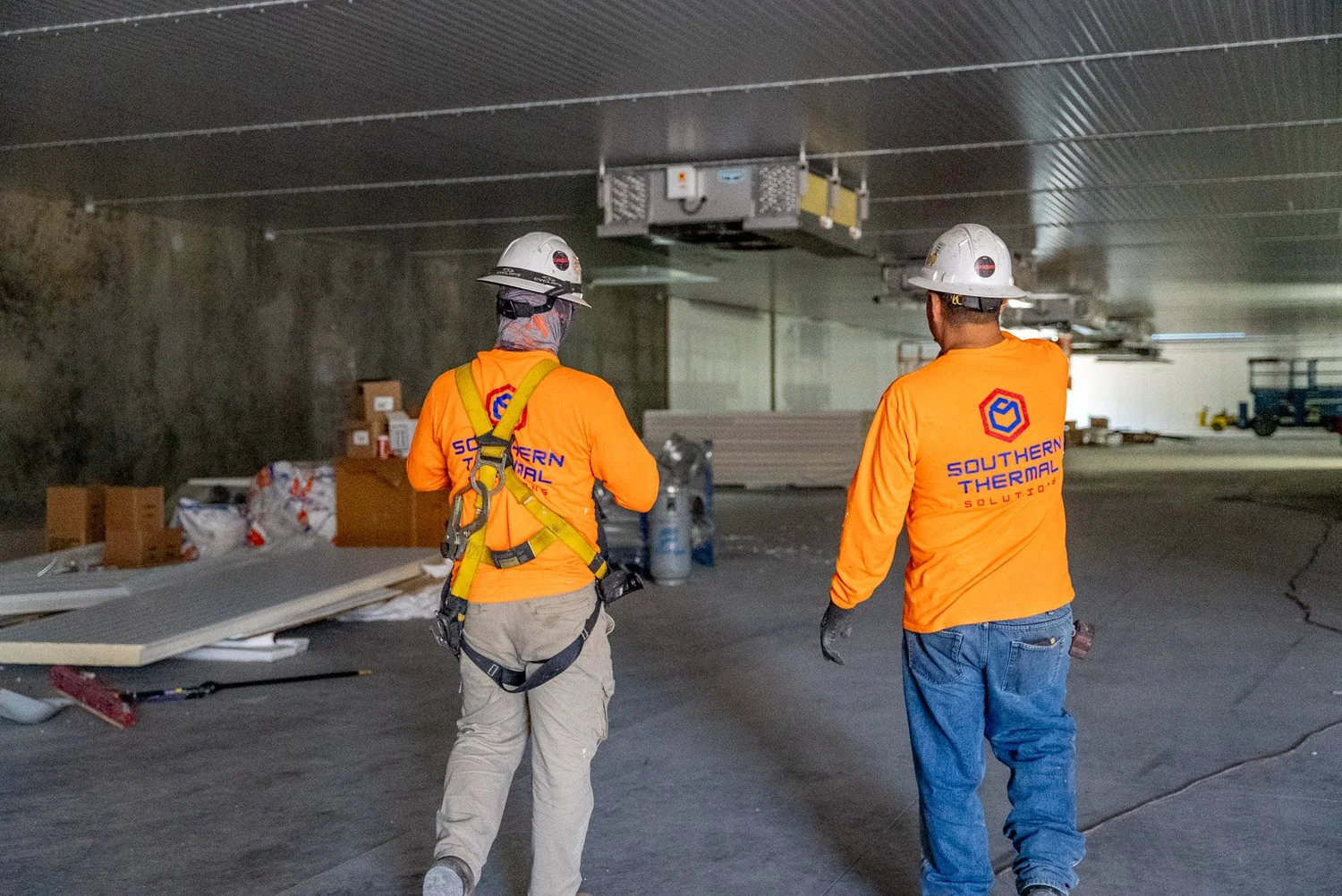 Two construction workers wearing orange shirts and white helmets walking through a dimly lit tunnel or underground construction site with stacks of materials and equipment in the background.