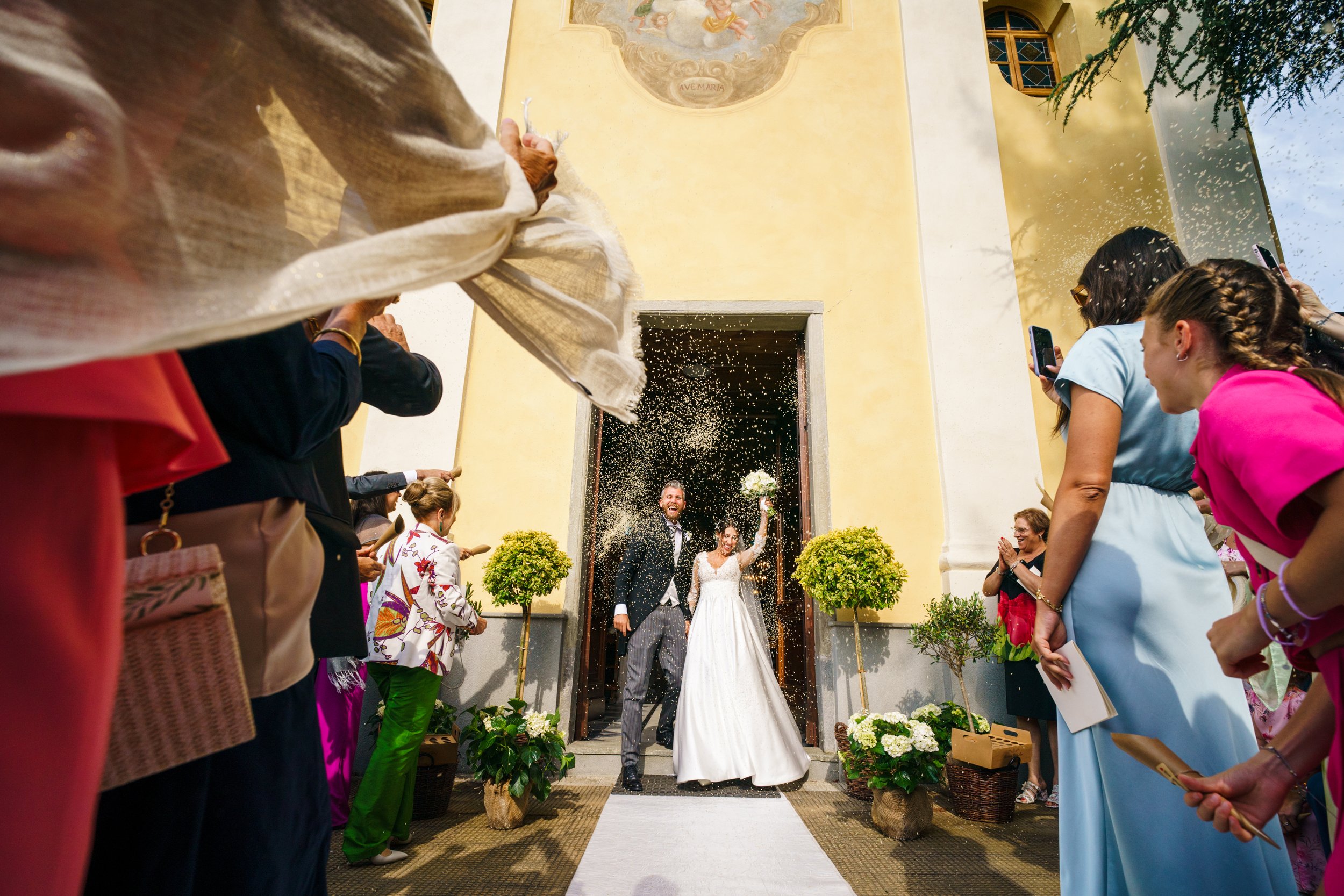 Sposi escono da una chiesa mentre vengono accolti da amici e familiari che lanciano riso, simbolo di buon augurio e prosperità. La sposa indossa un abito bianco e tiene in mano un bouquet. I presenti indossano abiti colorati e scattano foto con i tel