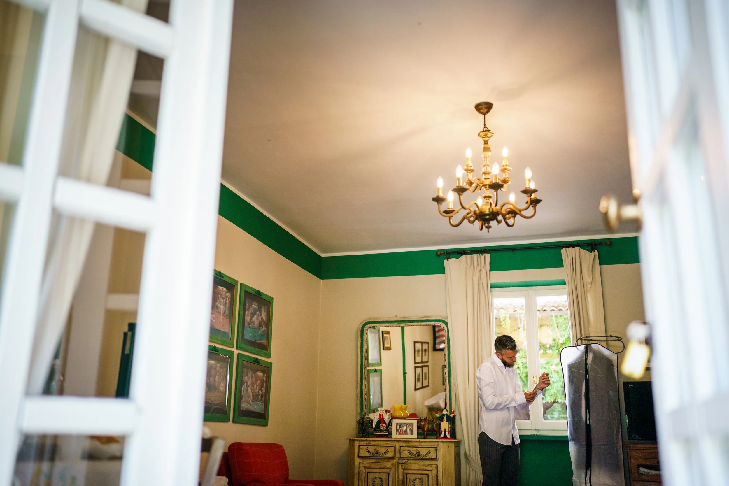 Interno di una stanza con un uomo vicino a una finestra, mentre si sistema il polsino della camicia. La stanza è arredata con un lampadario, quadri sulle pareti e un mobile con specchio. Usi colorati decorano la parete.