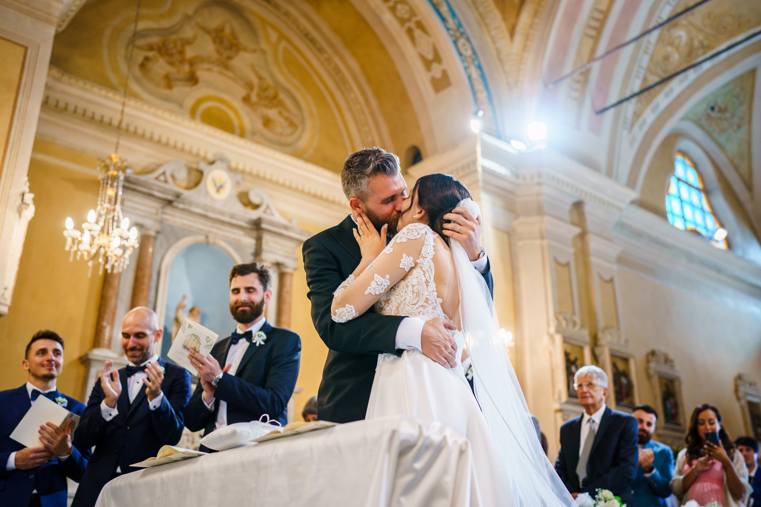 Una coppia di sposi si bacia in chiesa, circondata da invitati che applaudono. Gli interni della chiesa sono decorati con affreschi e candelabri.