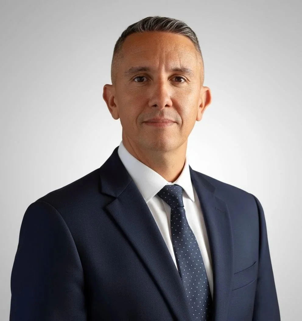 A professional man in a navy suit with a white shirt and a navy tie with white polka dots, standing against a plain light gray background.