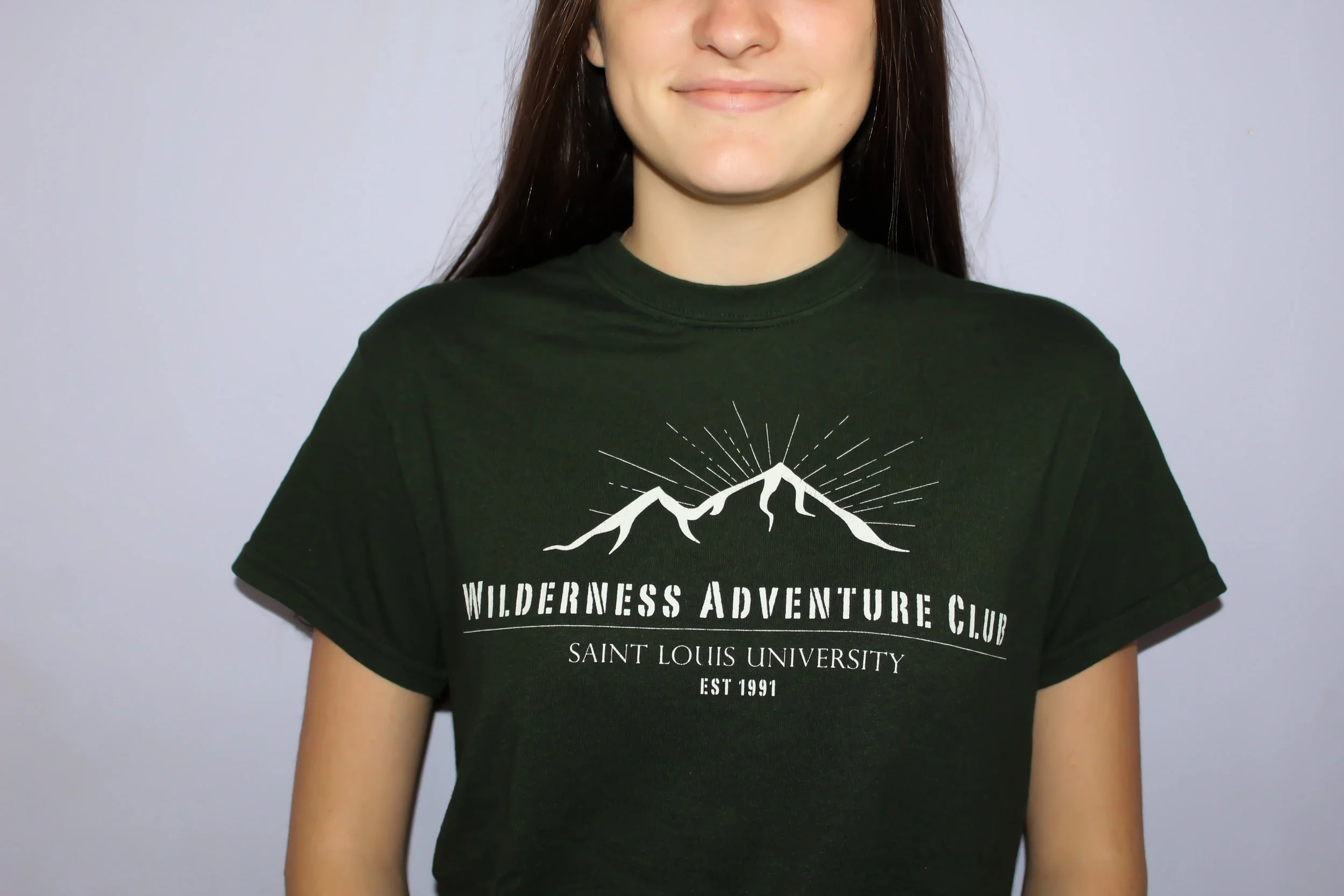 Close-up of a smiling woman wearing a black T-shirt with white mountain graphics and text that reads "Wilderness Adventure Club, Saint Louis University, EST 1991" designed and illustrated by Chicago-based medical artist and designer, Sophia Lappe.