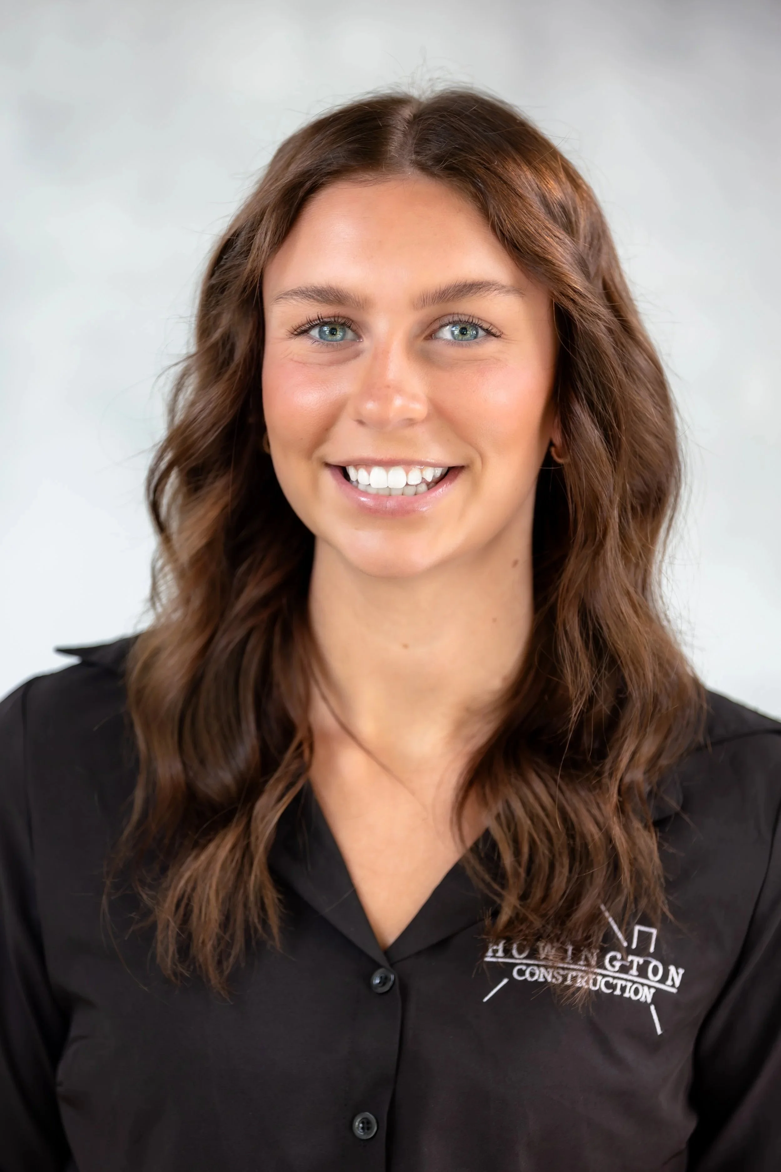 A young woman with long, wavy brown hair, smiling, wearing a black button-up shirt with a logo on it, black jeans, and a black belt with a gold G buckle, standing indoors against a light-colored wall.