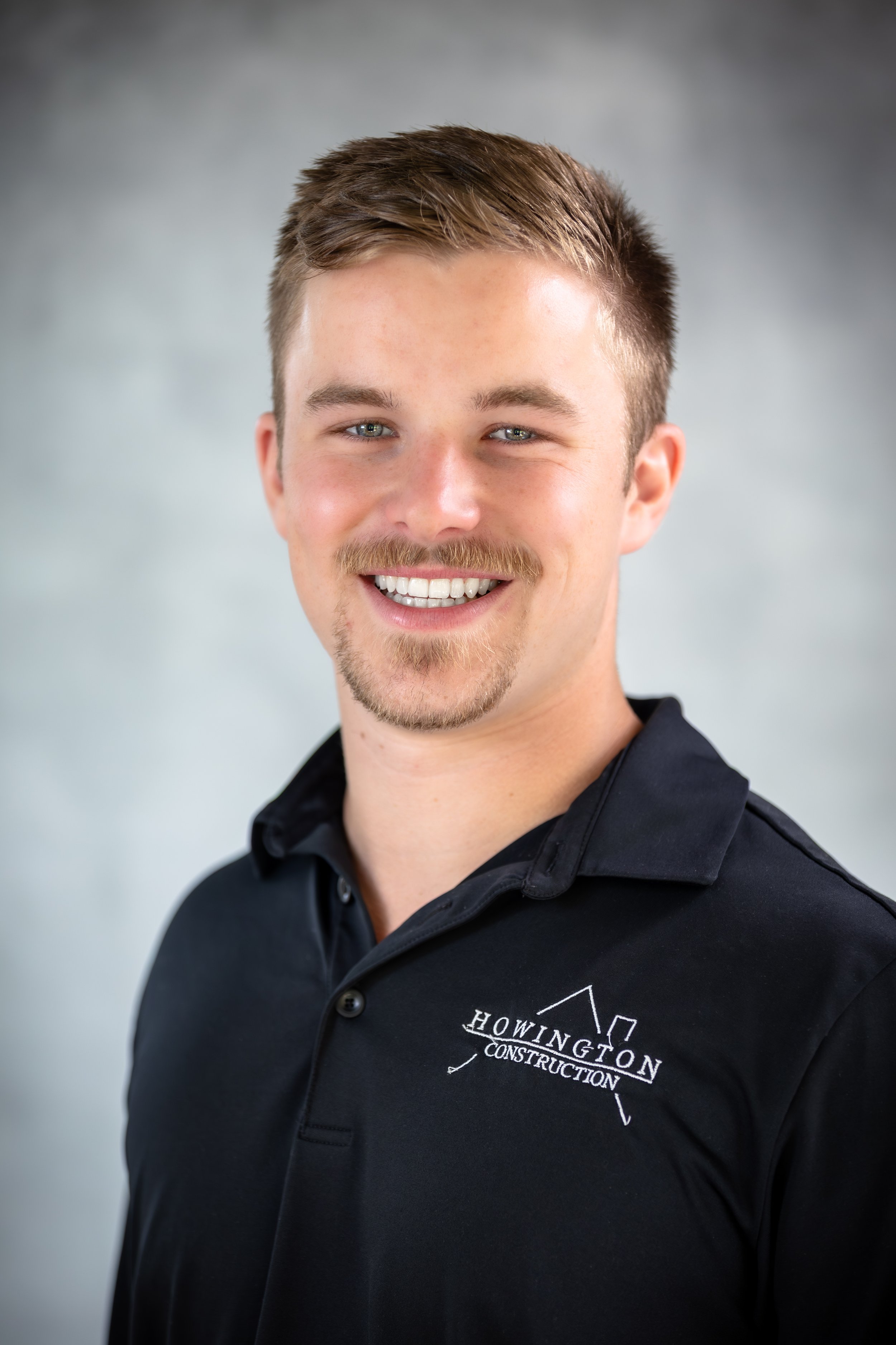 Young man smiling, wearing a black polo shirt with 'Howington Construction' logo, standing indoors with a white paneled wall background.