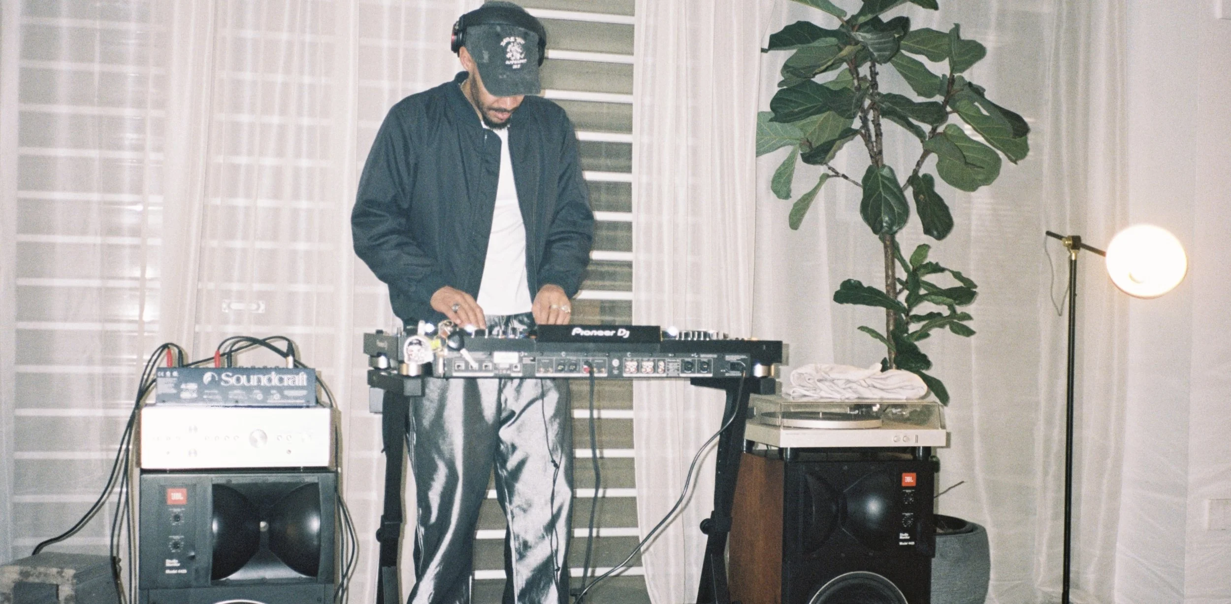 A man DJing in a living room, wearing a black jacket and patterned pants, with equipment including speakers, a mixer, and turntables in a room with curtains, a potted plant, and a floor lamp.