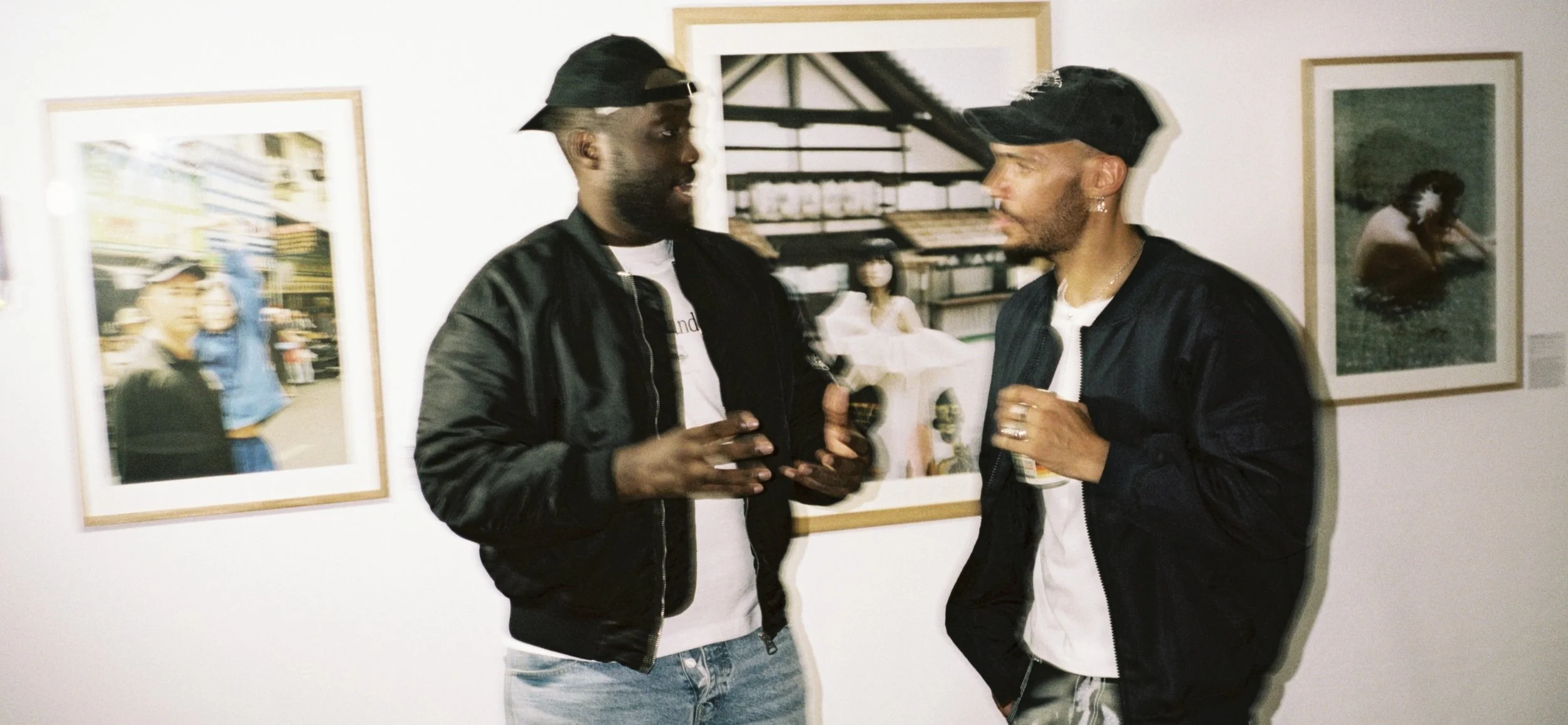 Two men are talking and holding drinks at an art gallery, with framed photographs hanging on a white wall behind them.