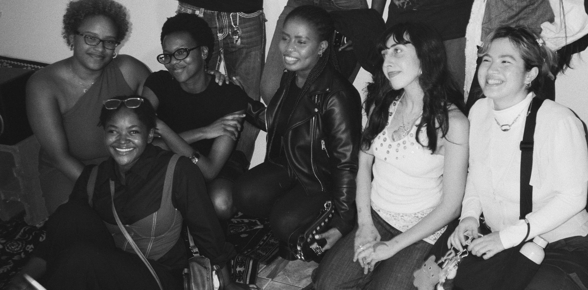 Group of six women, some wearing glasses, sitting and standing close together, smiling and enjoying each other's company in a social gathering.