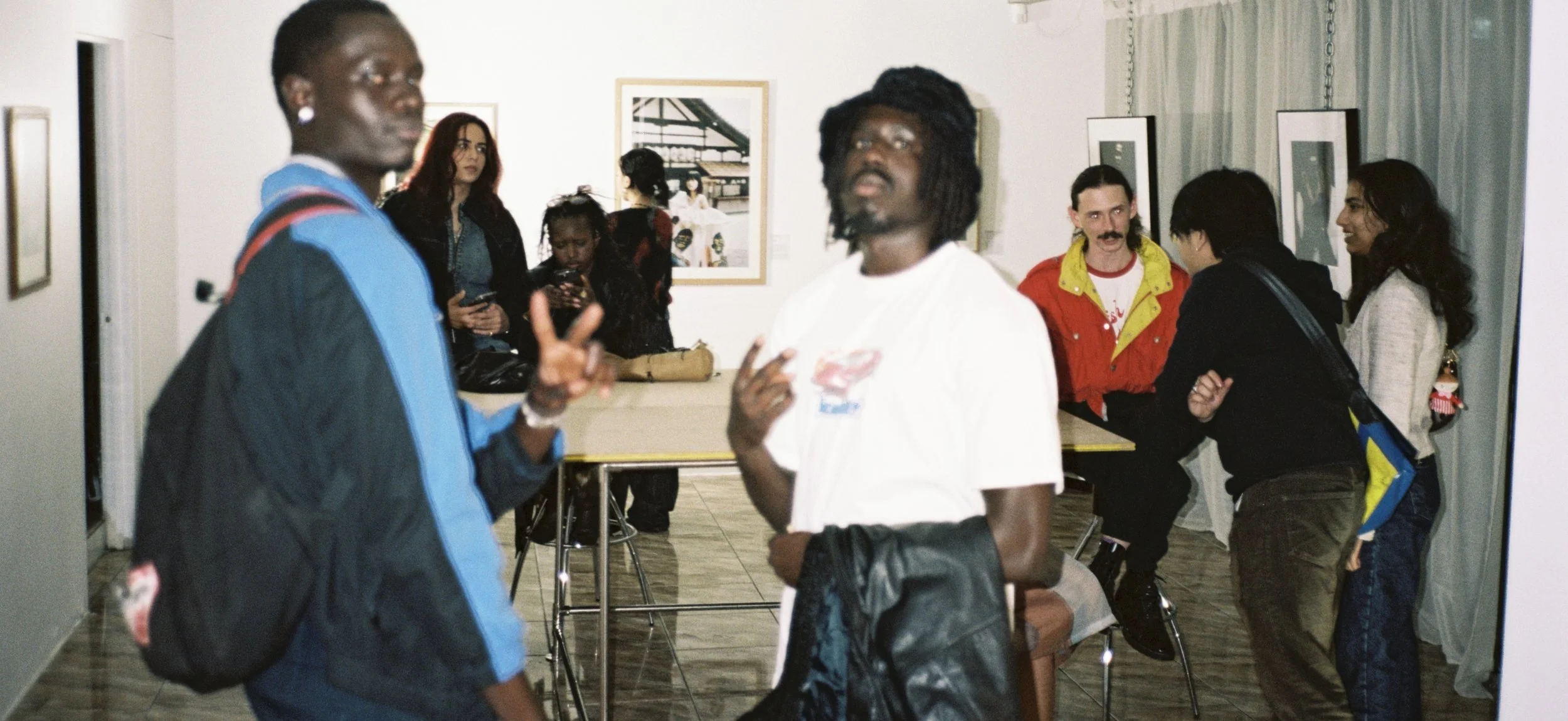 Group of people in an art gallery or exhibition space, some engaging in conversations, with framed artwork on the walls.